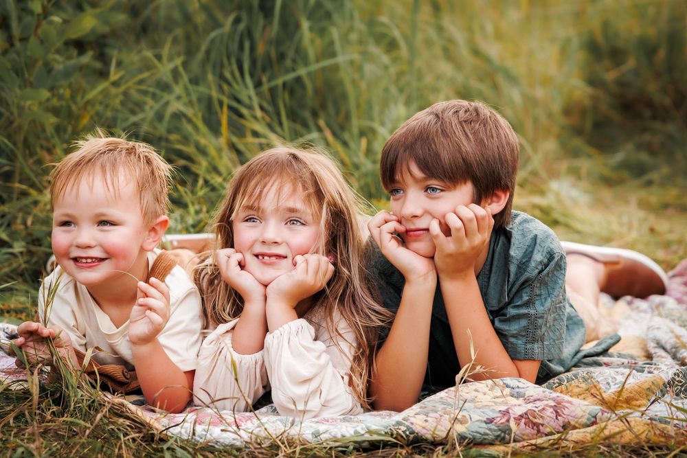 Three children lying together on a blanket in tall grass while smiling at the camera.