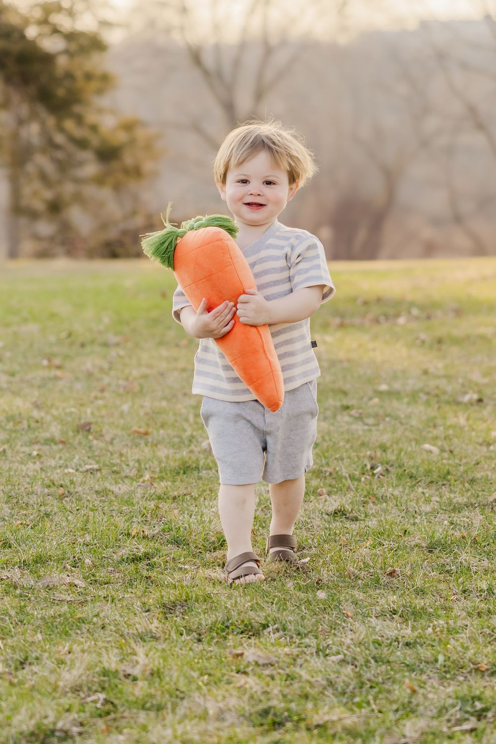 Small child in striped shirt holding large plush carrot toy outdoors.
