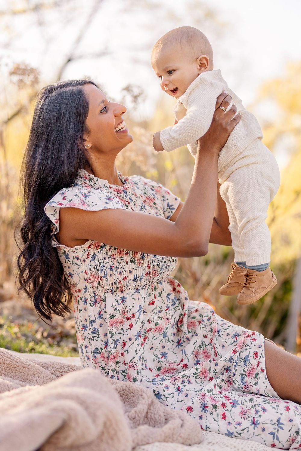 A heartwarming outdoor portrait of a woman in a floral dress holding up a smiling baby against golden sunlit scenery.