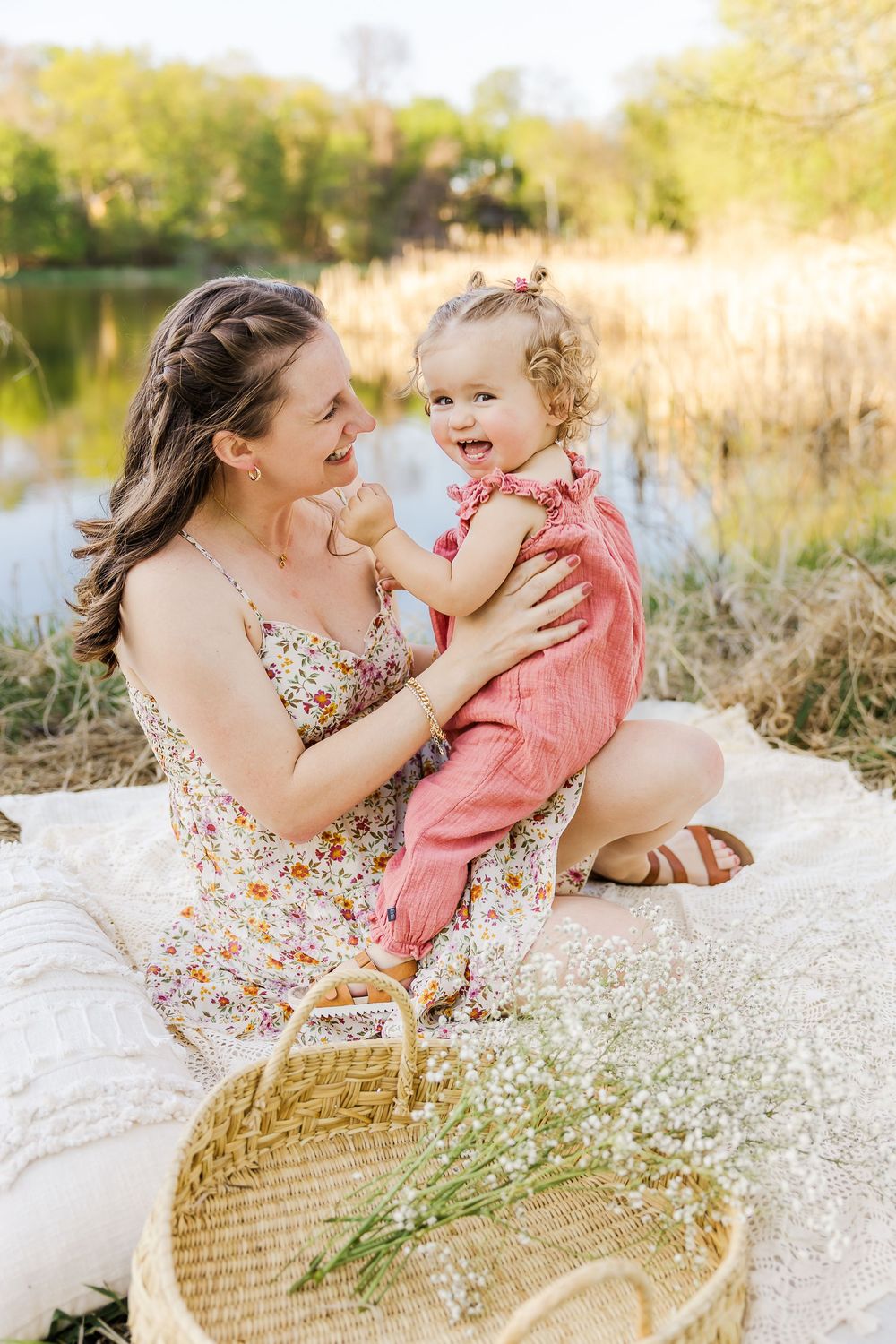 Mother and young child share joyful laughter while sitting on blanket near lake with straw basket and wildflowers.