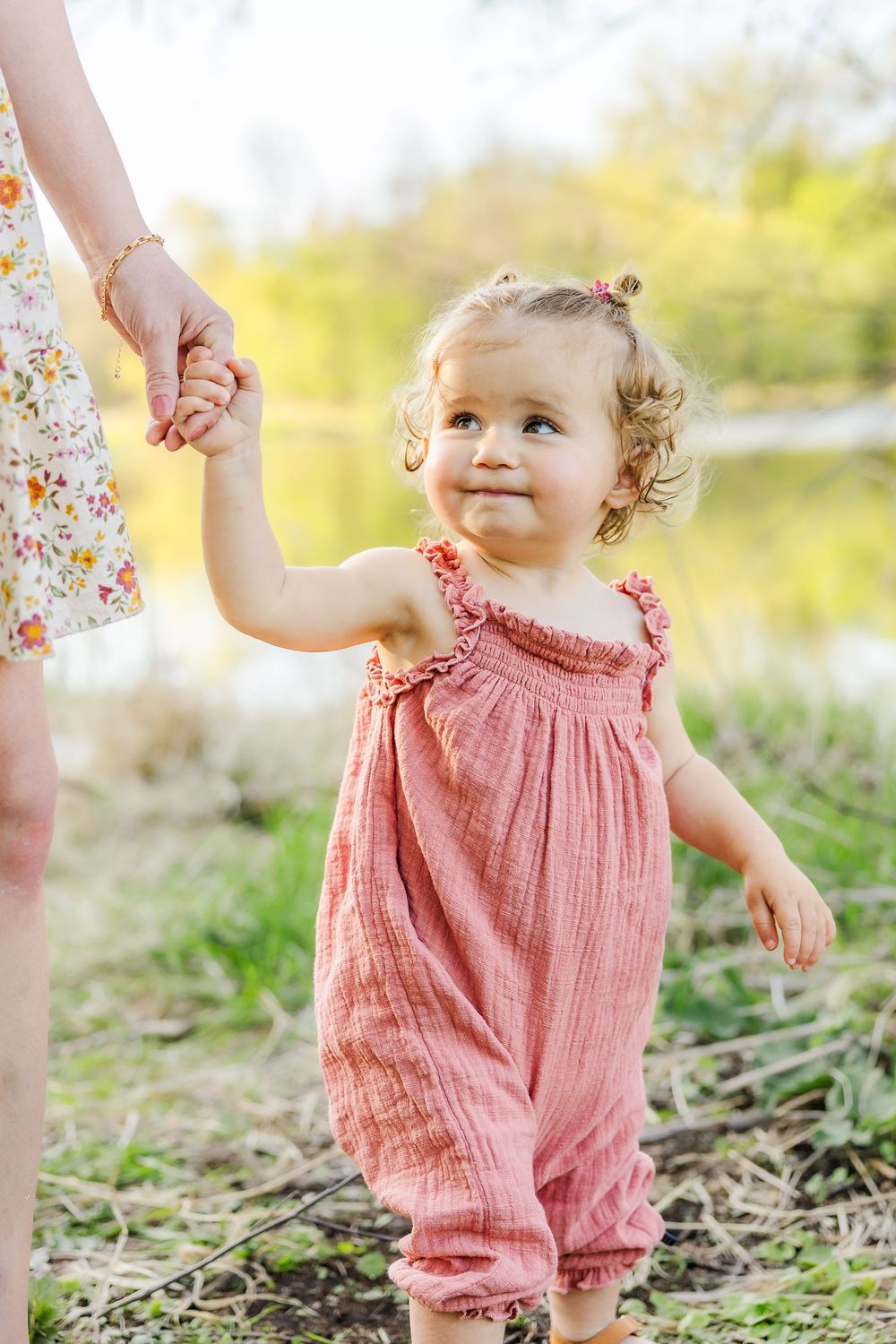 Young child in coral pink romper holds hands with parent while walking outdoors in springtime setting.