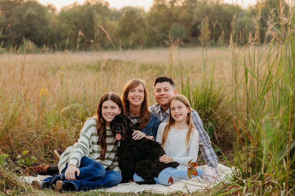 A group sits together on a blanket in a golden field during sunset surrounded by tall grass and autumn foliage.