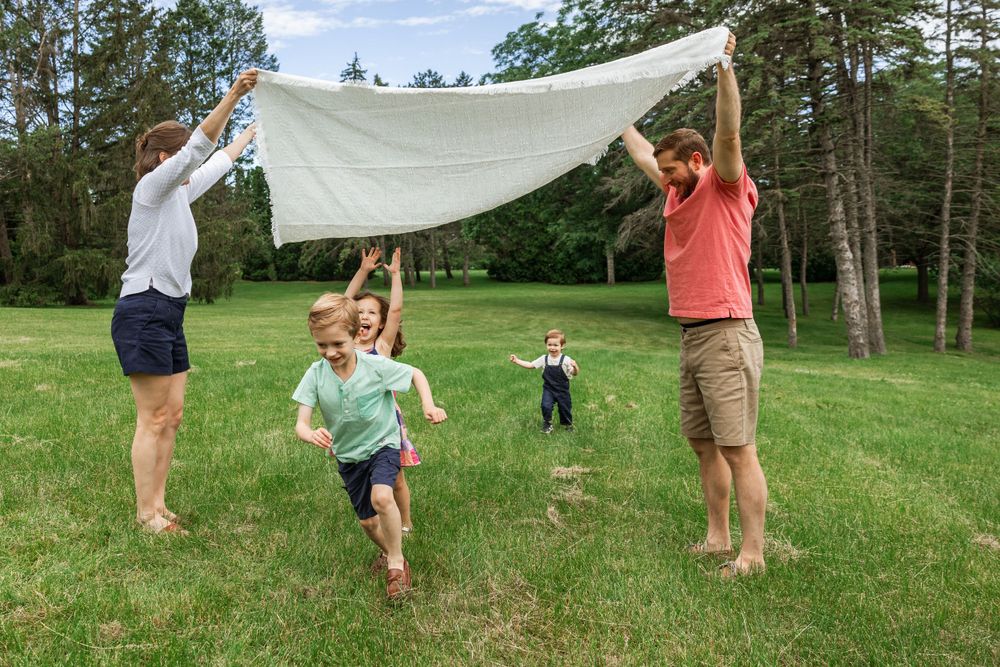 Family playing with white sheet parachute game in sunny green field.