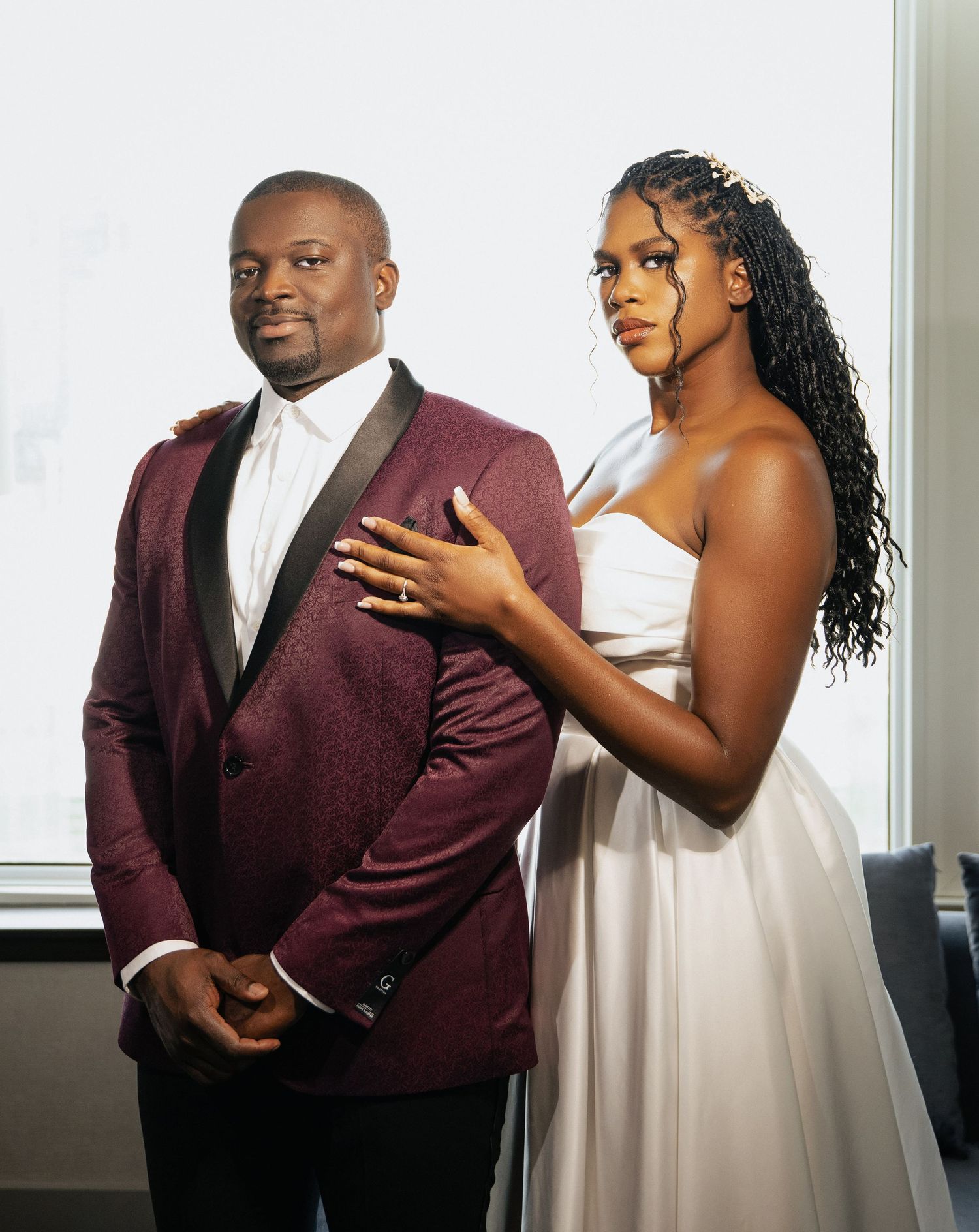 A couple poses in formal attire with the person wearing a burgundy tuxedo jacket and their partner in a white dress.