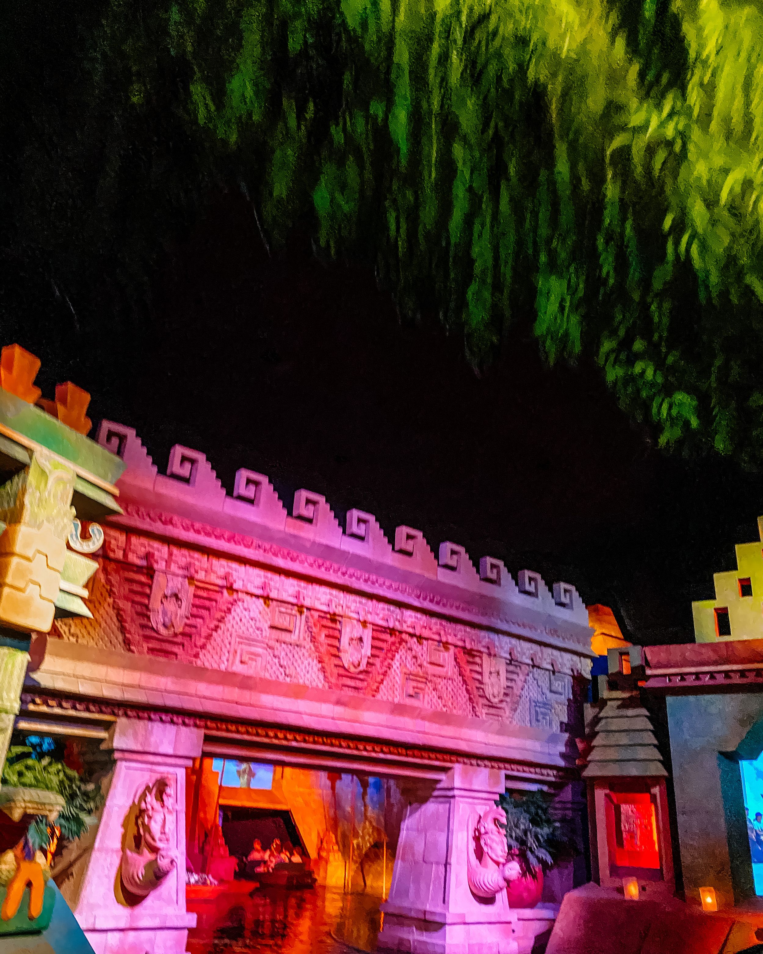 Ancient temple-style wall facade illuminated with pink and colorful lighting against dark foliage at night.