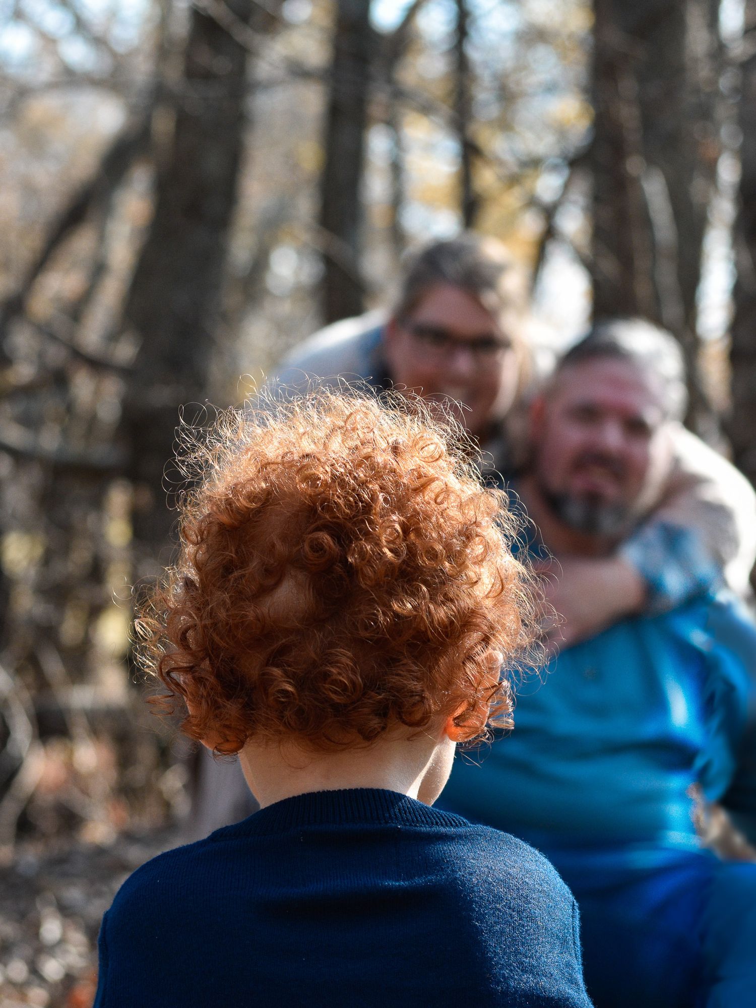 Child with curly red hair in the foreground looking at two adults in the background, blurred, standing in a wooded area.