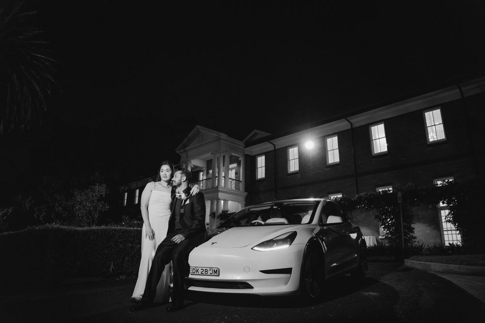 Nighttime wedding exit photo with a Tesla car parked outside a lit building.