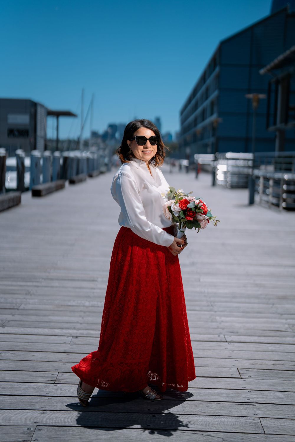 A figure in a white blouse and red skirt holds a bouquet on a wooden boardwalk by a marina.
