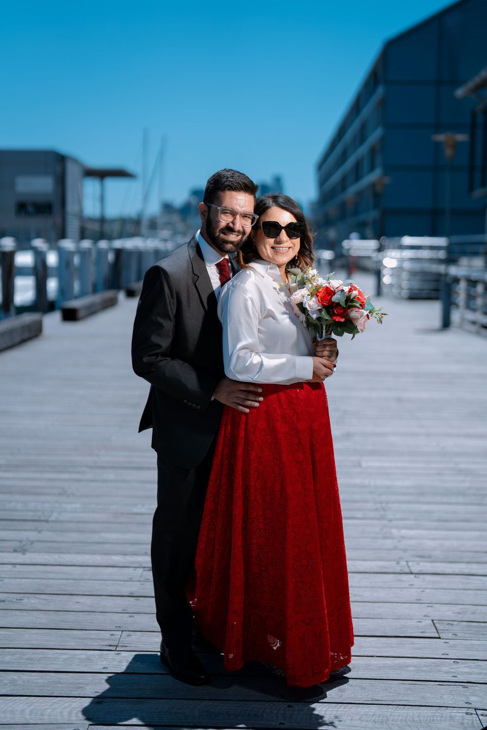 A couple poses on a marina boardwalk, with the woman in a red skirt holding a floral bouquet.