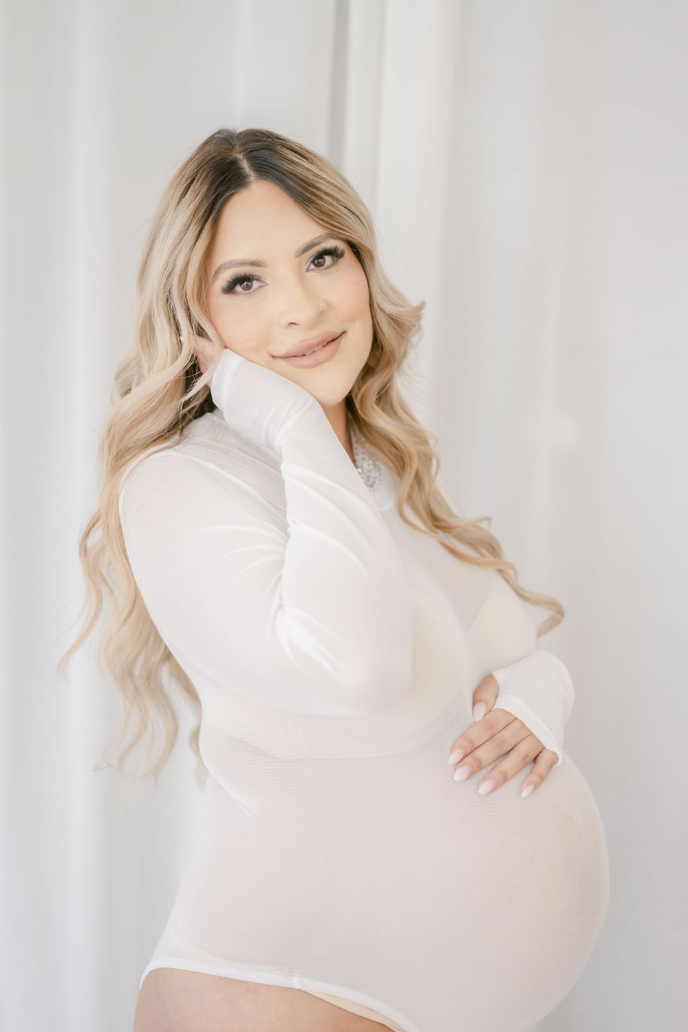Maternity portrait of a woman in a white turtleneck sweater posing against a light backdrop.