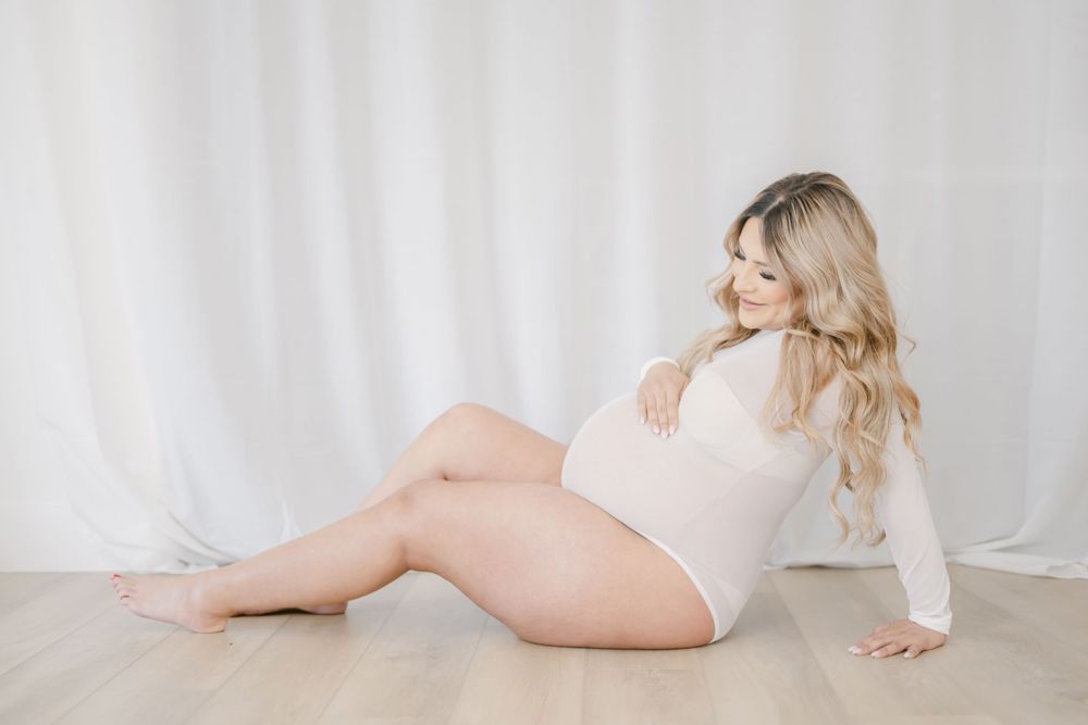 Woman in soft white bodysuit poses elegantly on floor against light backdrop in intimate portrait session.