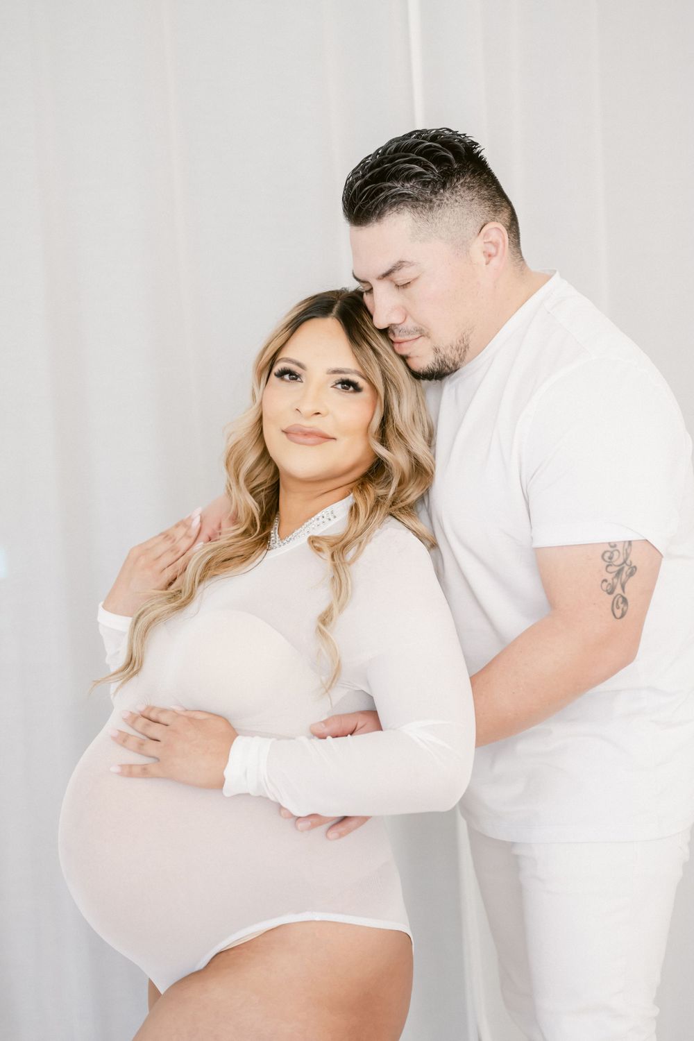 Intimate maternity photo features couple in white attire embracing against light background during pregnancy photoshoot.