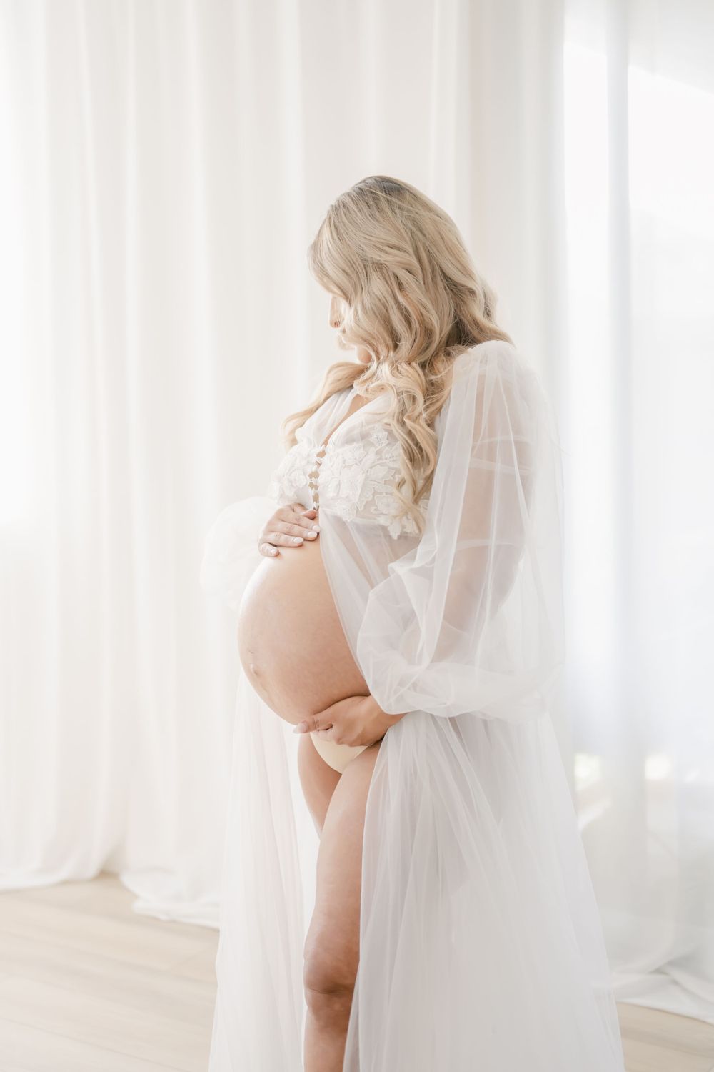 Ethereal maternity photo features an expectant mother in a flowing white robe and lingerie against sheer curtains.