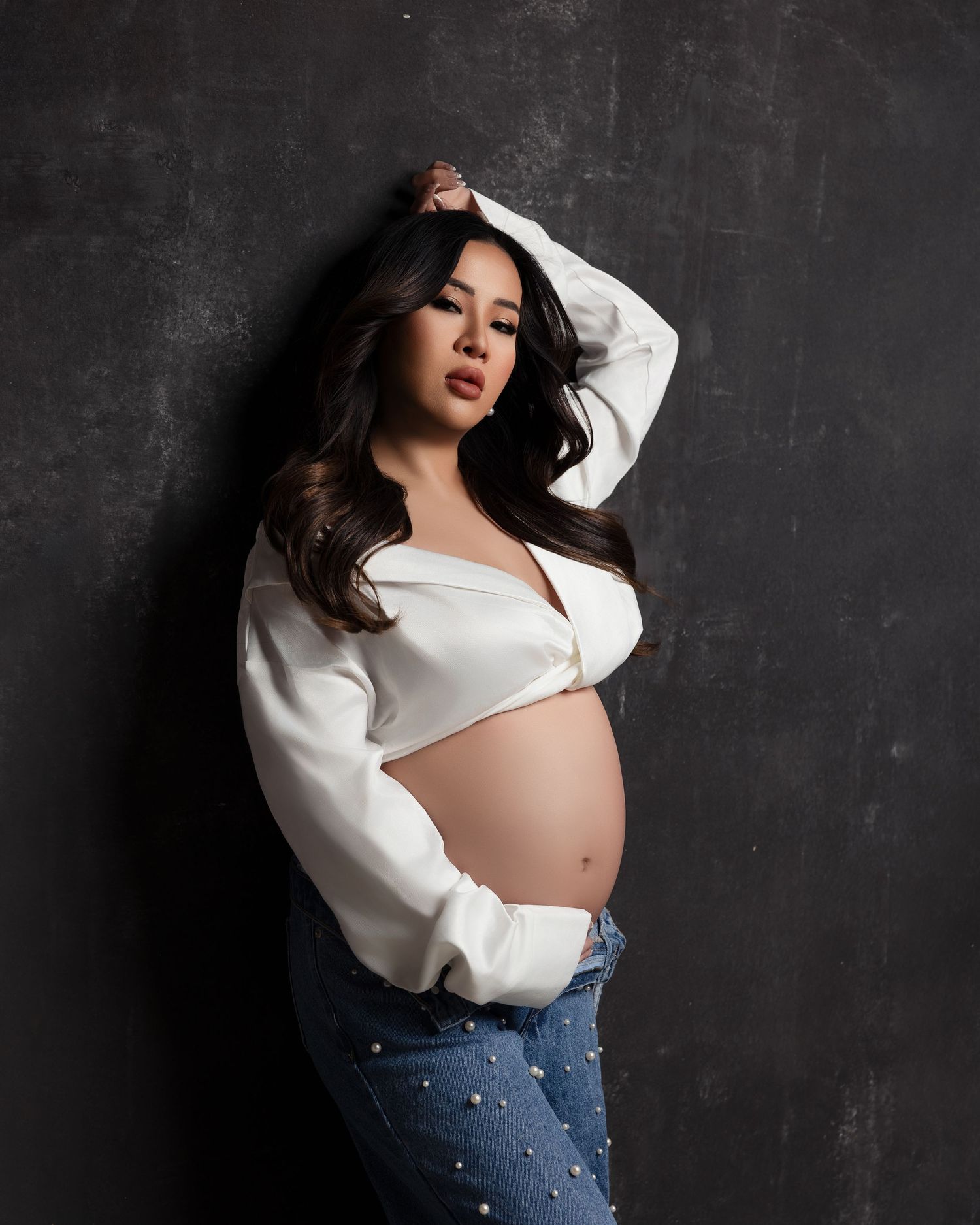 A pregnant woman in a white sweater and embellished jeans poses against a dark textured wall.