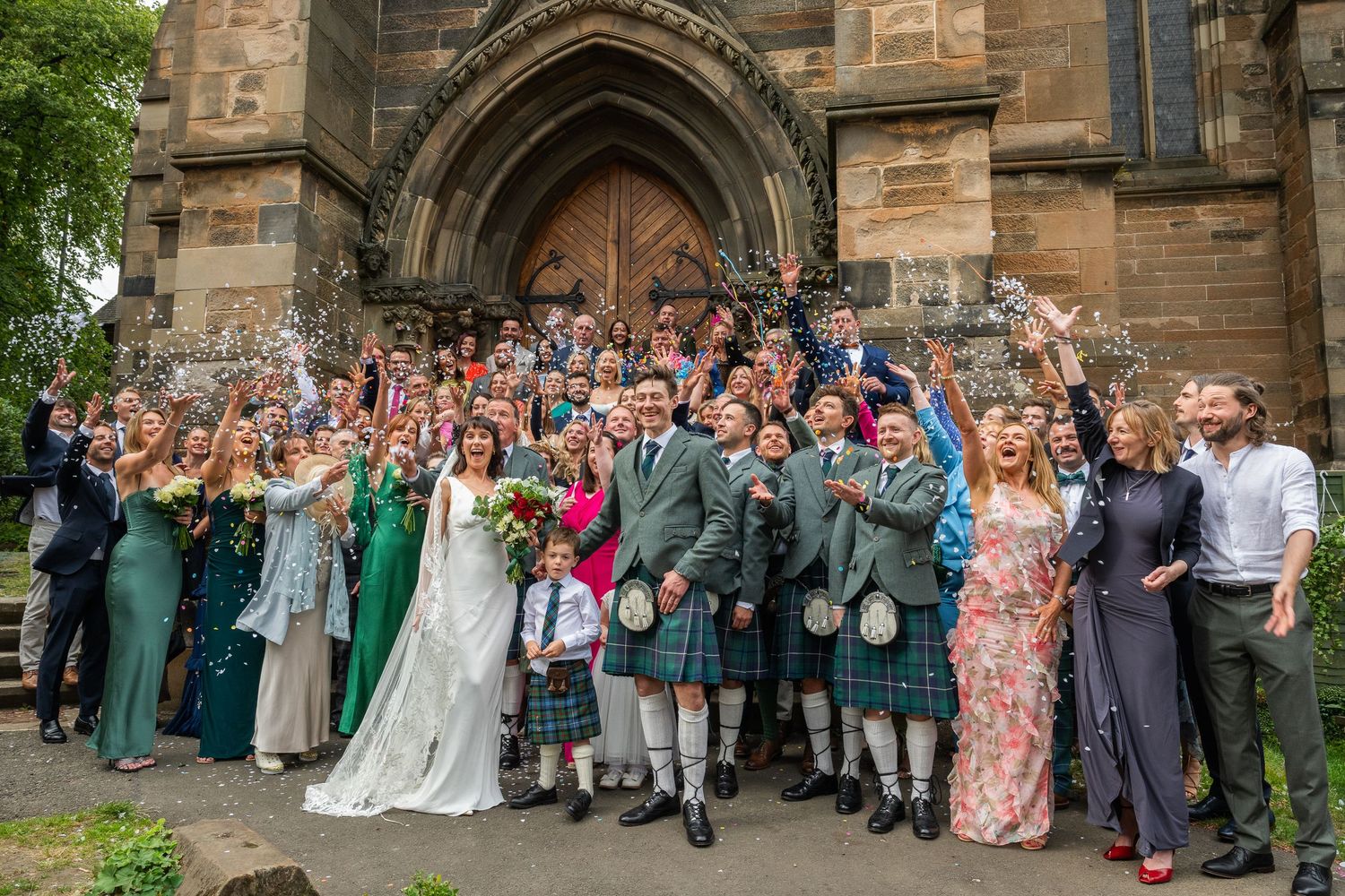 Large group wedding photo outside historic stone church with gothic architecture.