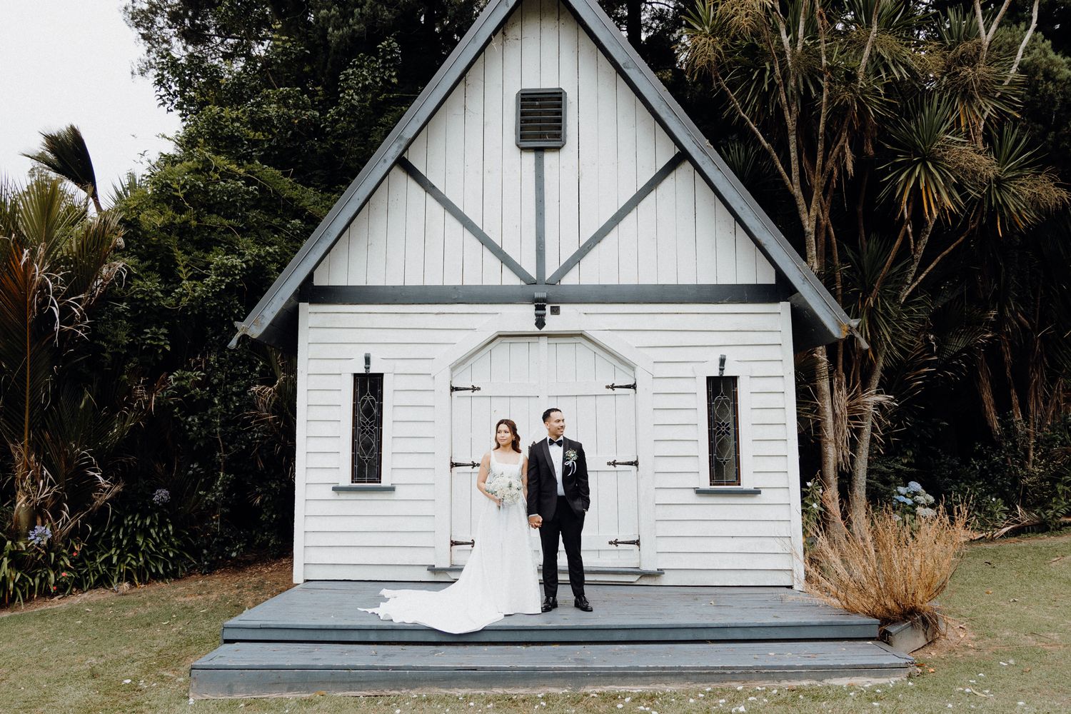 A couple stands together in formal attire in front of a charming white wooden chapel captured by Apertured Media Auckland Wedding Photograph