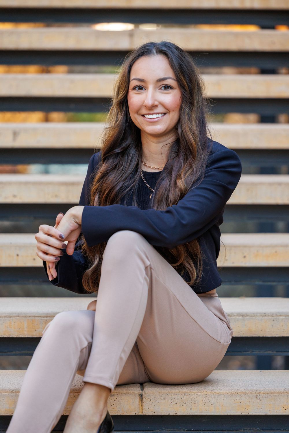 Businesswoman in black blazer and khaki pants sitting casually on wooden steps outdoors.