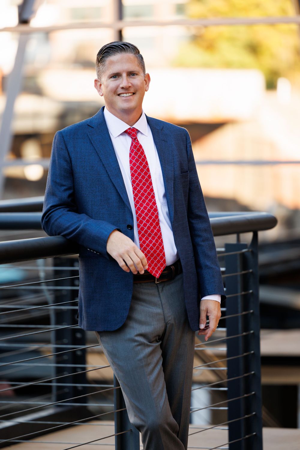 Business professional in navy blazer and red tie poses outdoors against modern architecture.