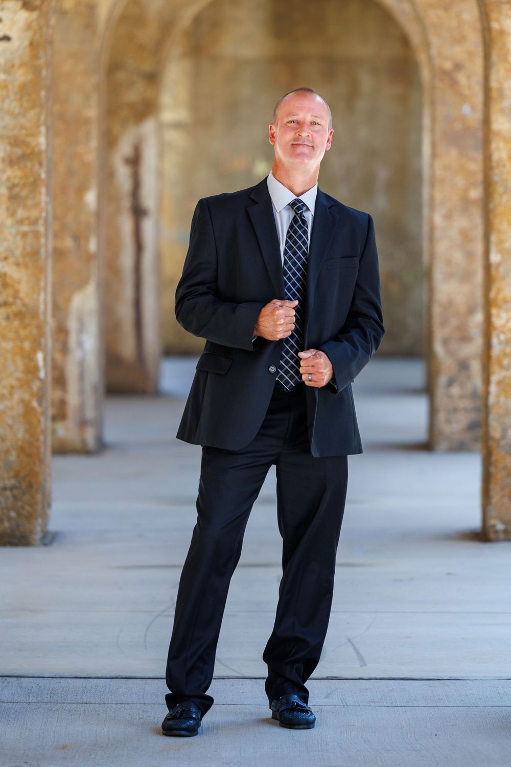 Business professional in dark suit and tie standing in arched stone corridor.