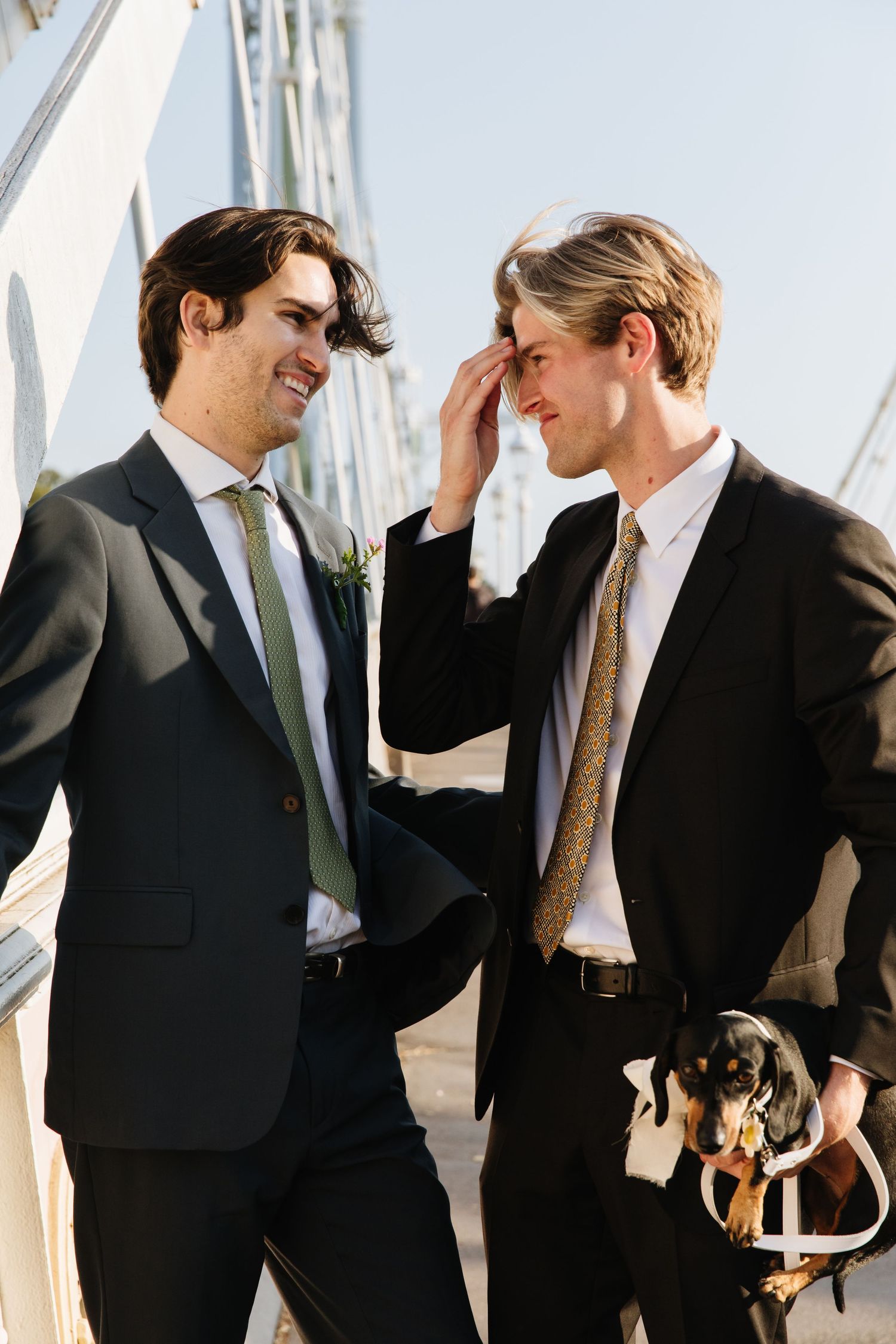 Two people in suits share a lighthearted moment outdoors on a sunny day.