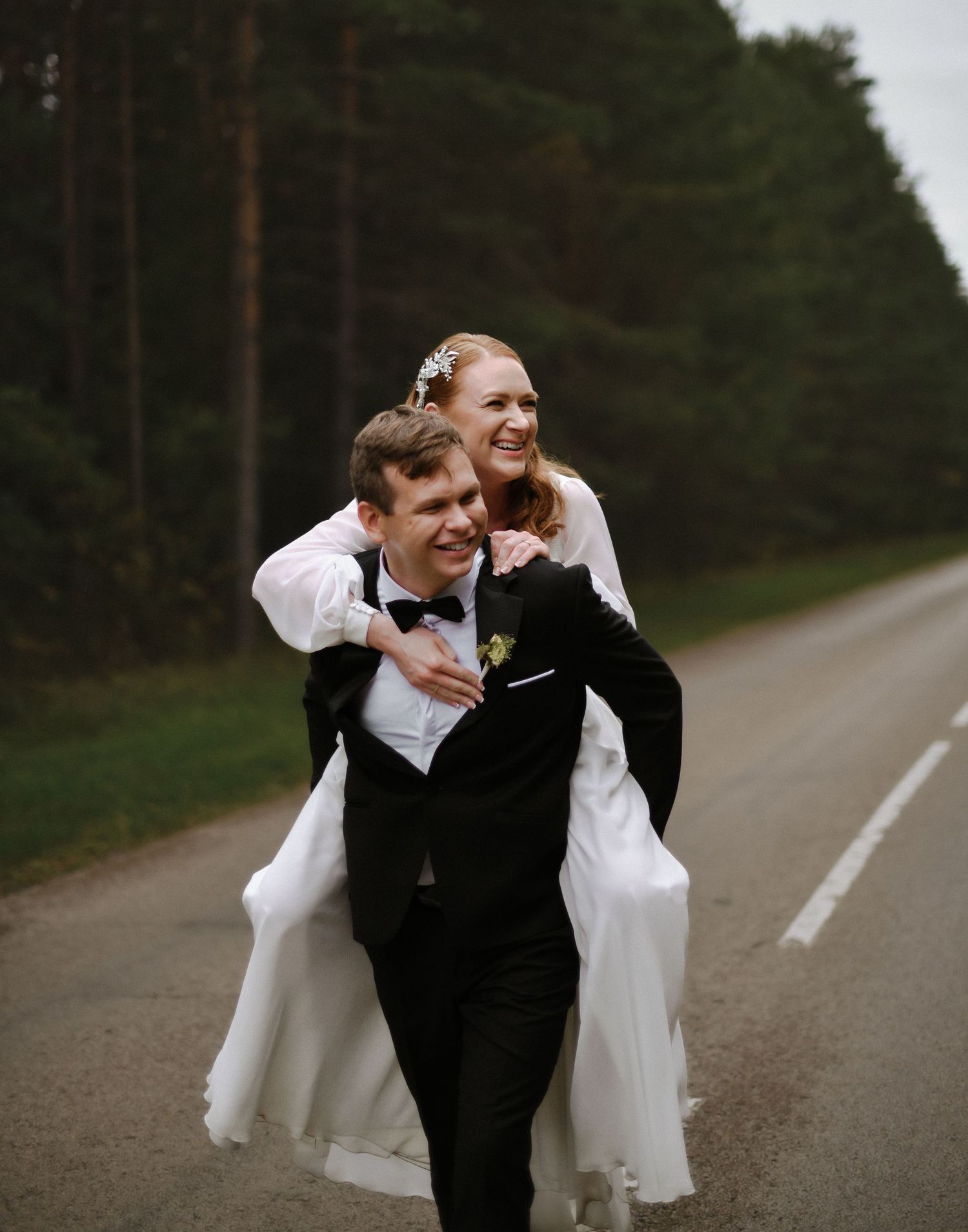 Groom giving bride a piggyback ride on a rural road surrounded by pine trees.