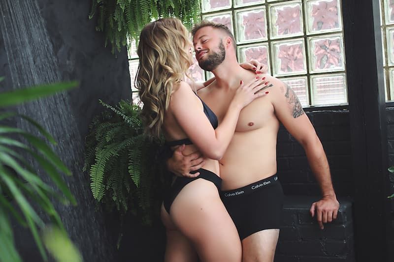 An intimate couple in black underwear embraces near a window with ferns and plants in the foreground.