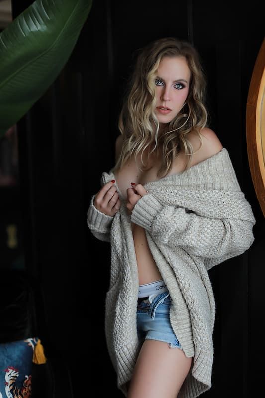 Model in oversized knit cardigan and denim shorts poses against dark background with tropical plant leaves.
