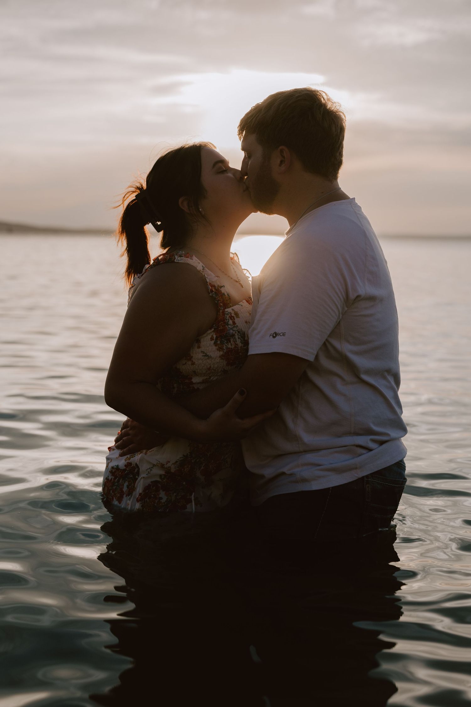 A romantic couple shares a sunset kiss while standing together in the ocean waters at dusk.