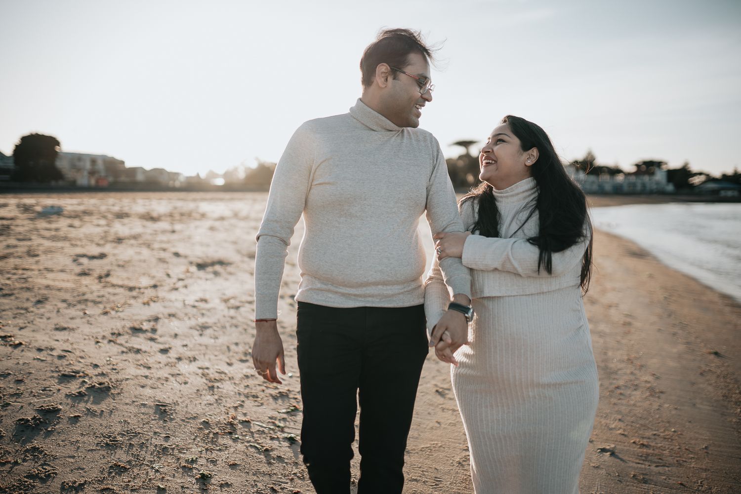 Couple dressed in warm ivory sweaters walks together on a beach at sunset, sharing a tender moment taken by Melbourne maternity photographer