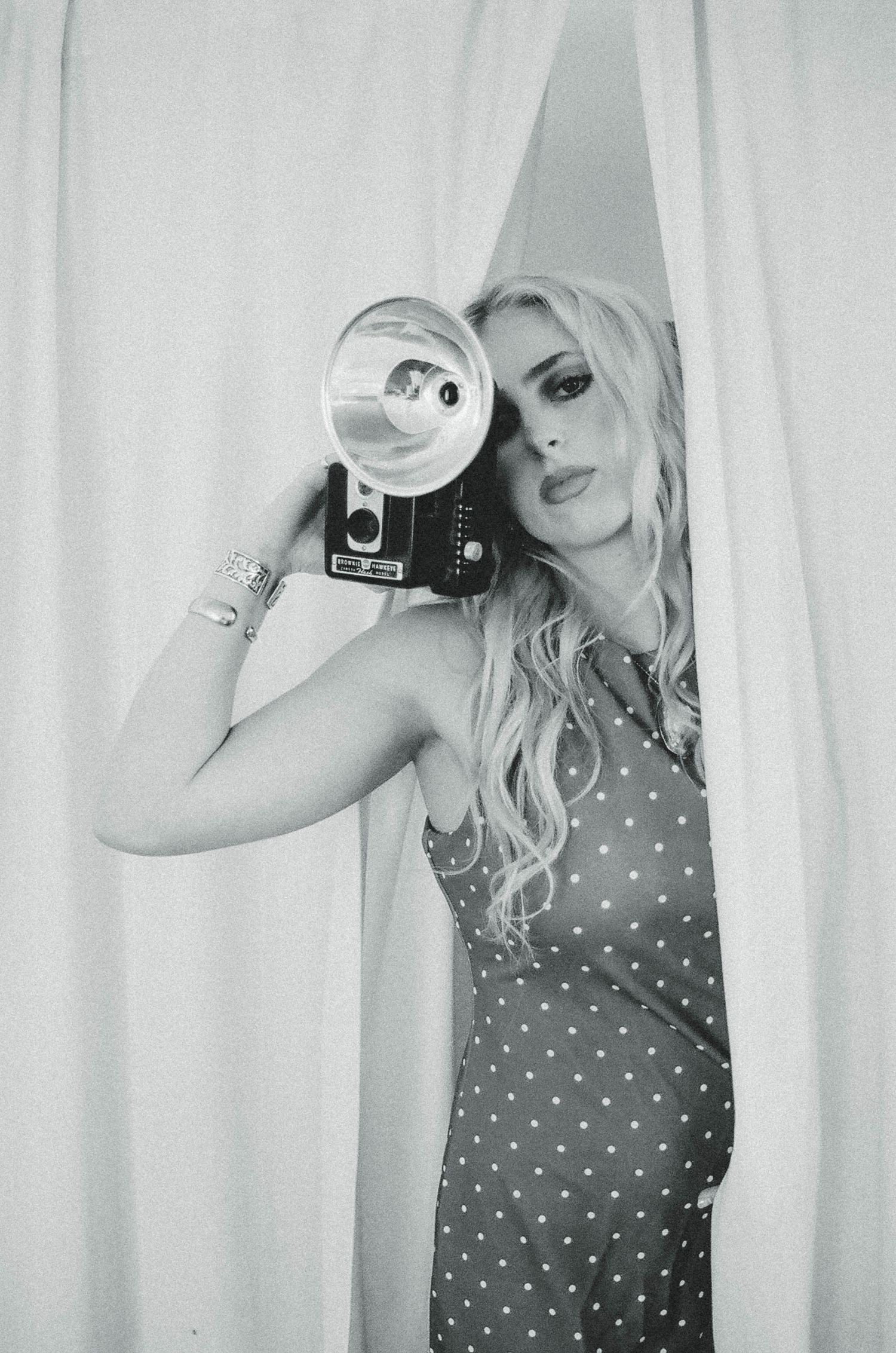 Person in denim dress and white cardigan holding vintage camera while sitting outdoors in natural lighting.