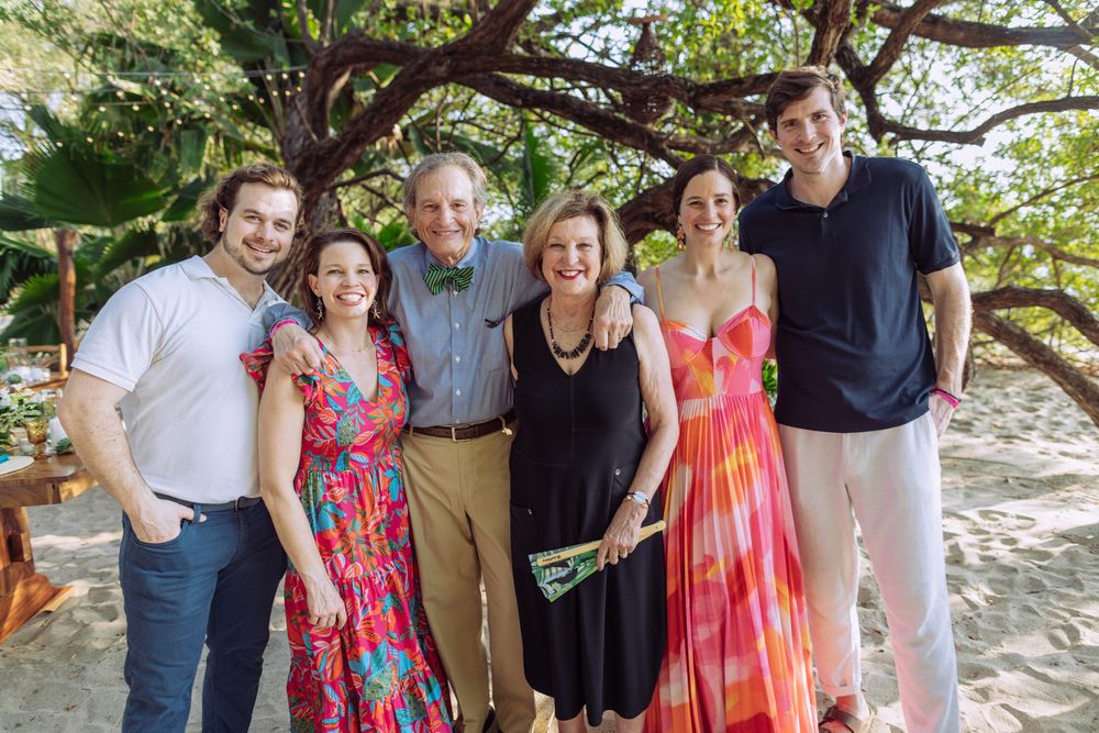 A group stands together outdoors under a large tree branch on a sunny day wearing colorful summer attire.