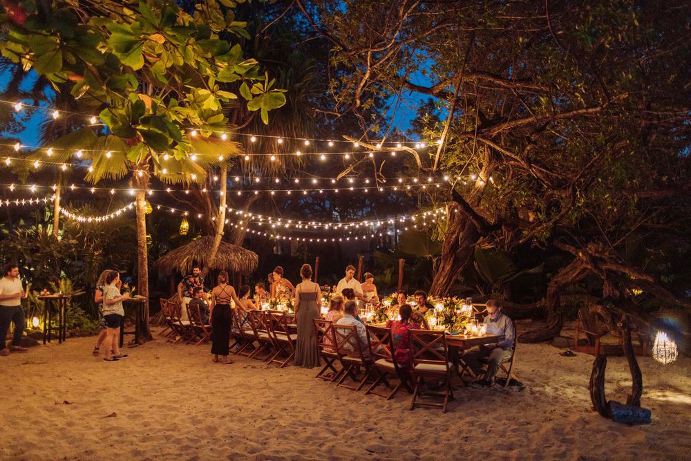 Romantic outdoor dinner party at night with string lights illuminating long tables on a sandy beach setting.