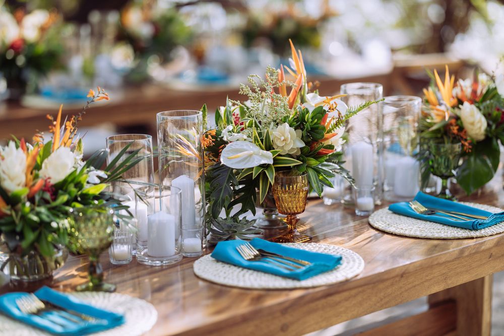 Elegant table setting with tropical flowers, teal napkins and vintage glassware on wooden farmhouse table.