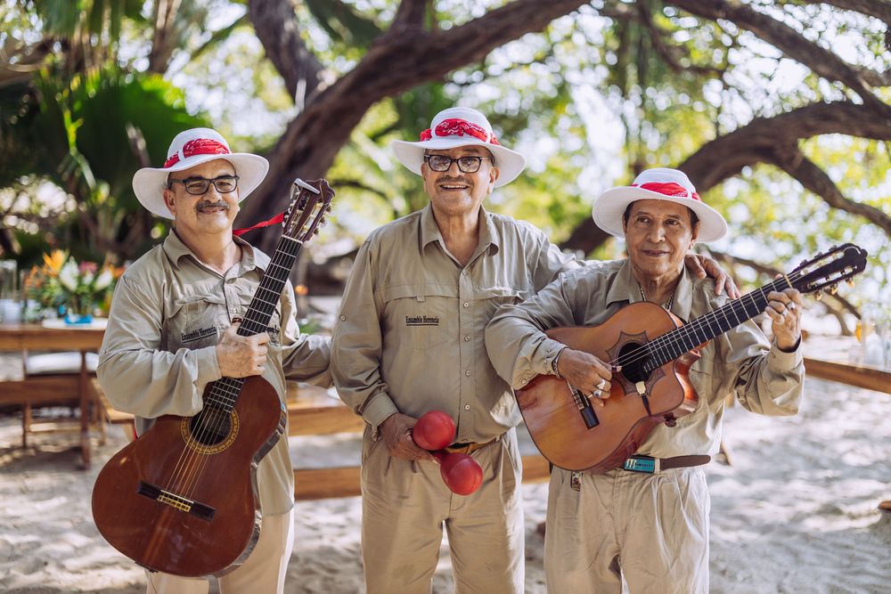Three musicians wearing Santa hats and beige outfits pose with acoustic guitars under a tree on a sunny day.