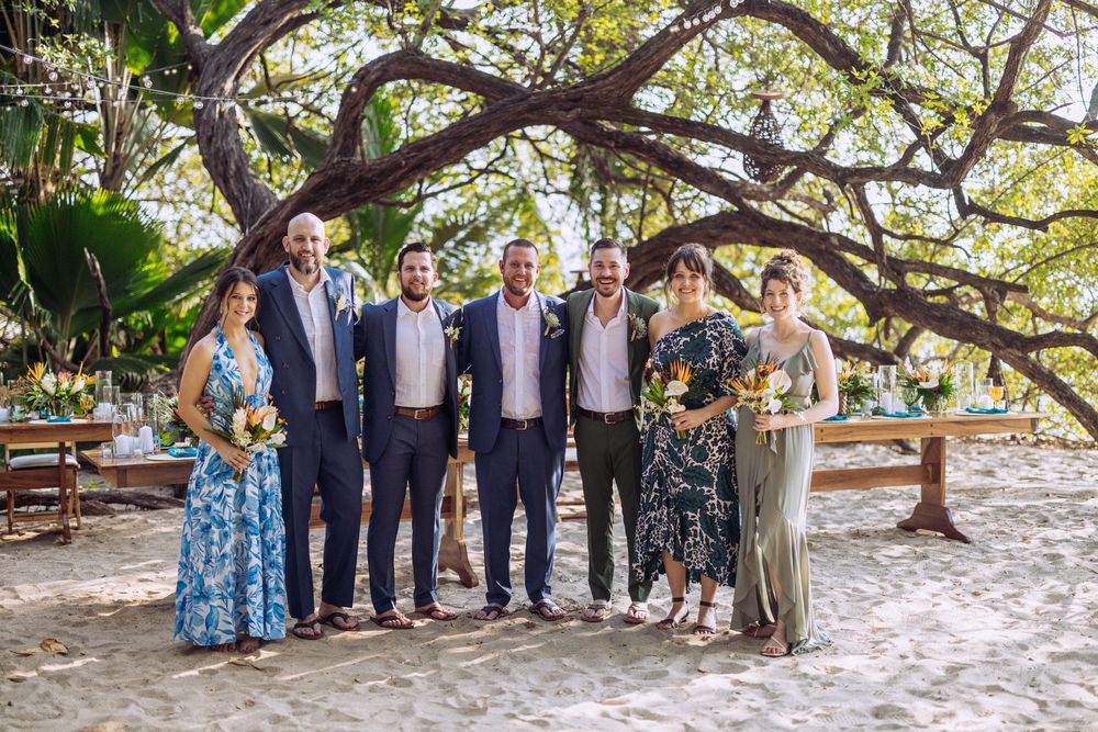 Wedding party in formal attire standing together under arching tree branches at outdoor beach venue.