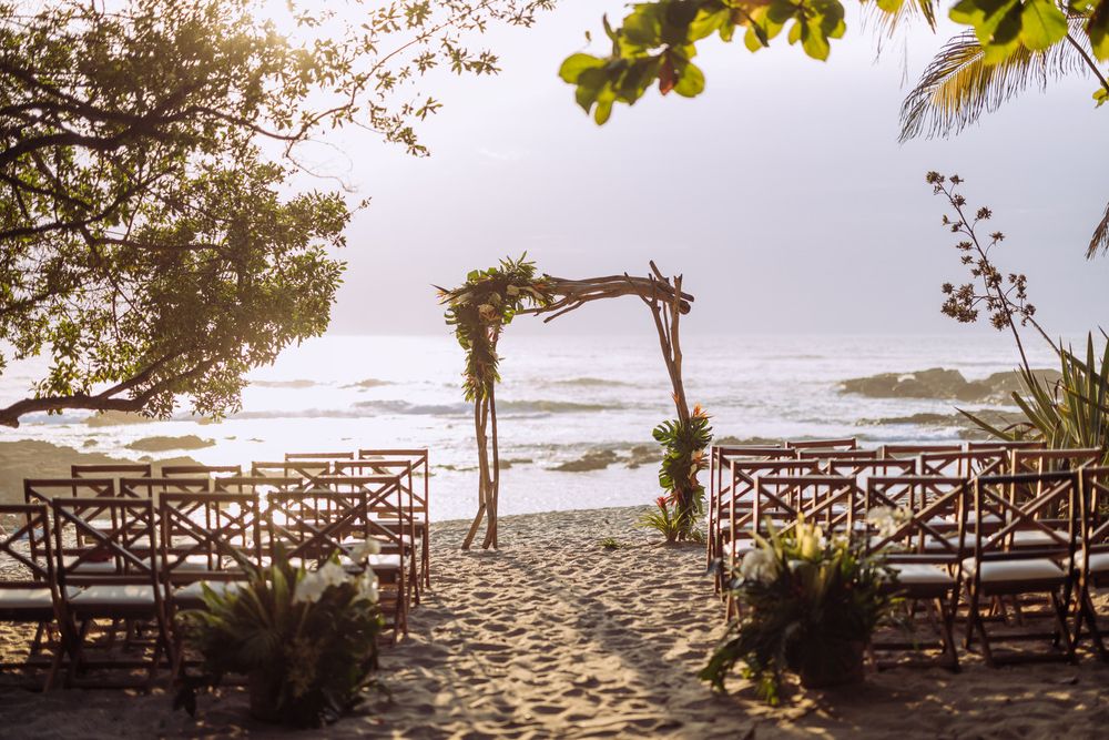 Beach wedding ceremony setup with wooden chairs and rustic arch overlooking the ocean at sunset.
