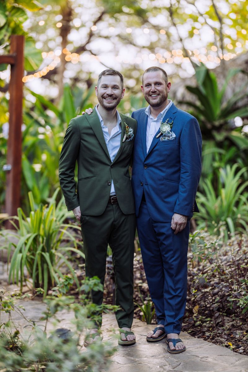 Two people pose together on a garden path wearing suits, one in green and one in navy blue with floral boutonnieres.
