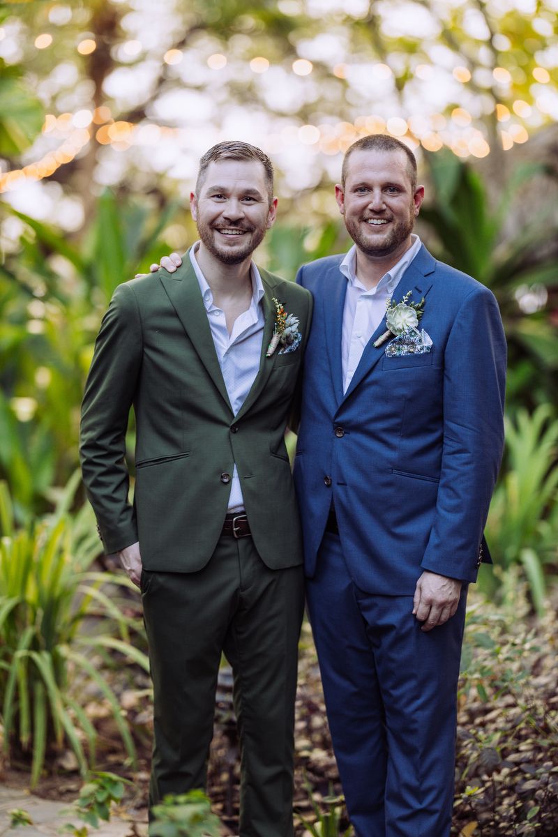 Two grooms in complementary suits of blue and green with floral boutonnieres stand together in a garden setting.