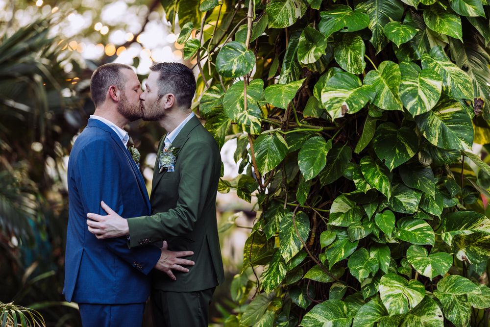 Two people in suits share an intimate moment against a backdrop of lush green foliage.