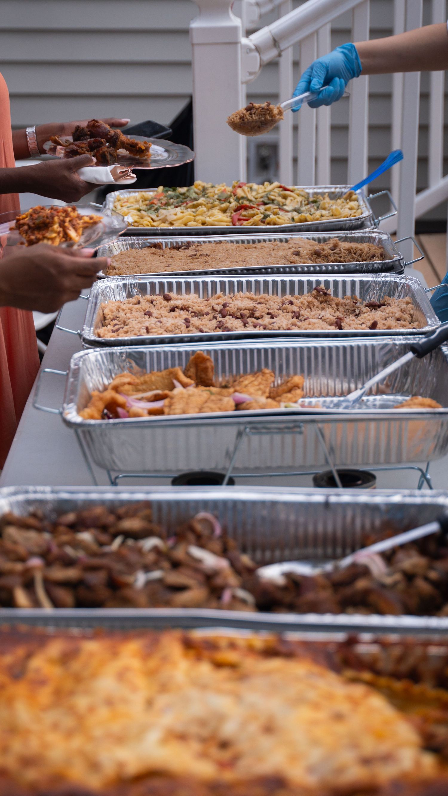 Buffet line of aluminum catering trays filled with various dishes being served outdoors on a deck or patio.