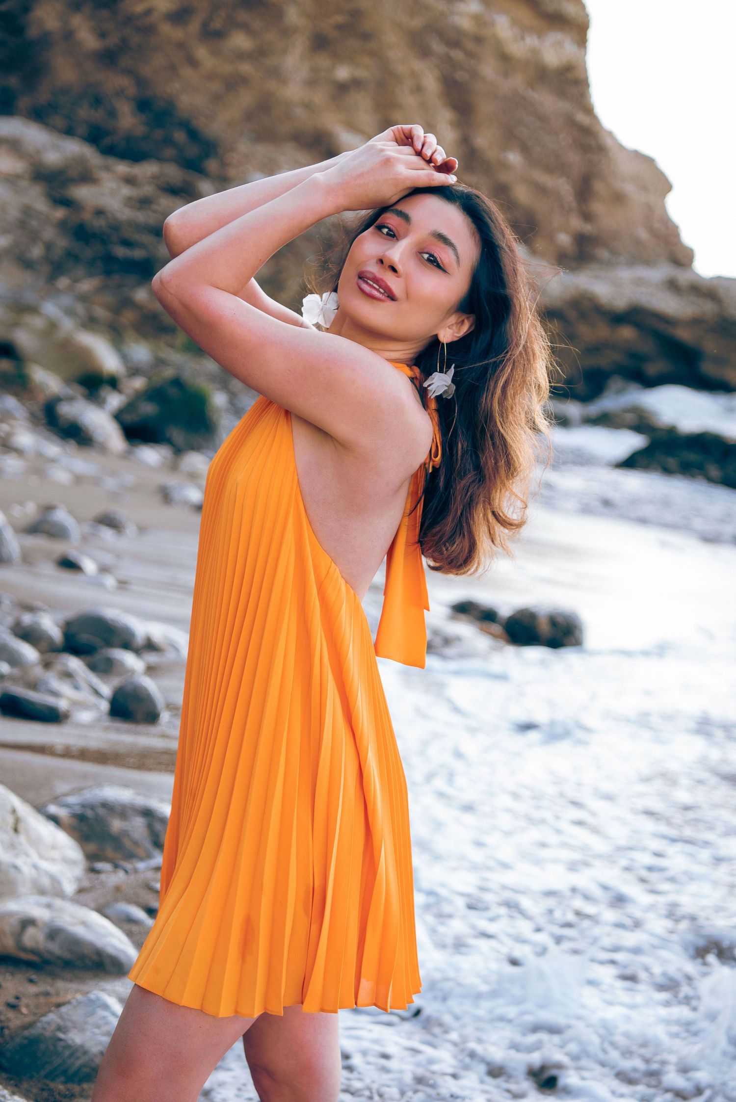 A bright orange pleated sundress flows in the breeze as someone poses elegantly on a rocky beach coastline.