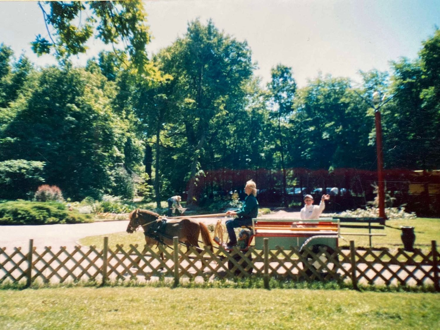 Horses and riders trot along a white lattice fence at an outdoor riding arena on a sunny summer day.
