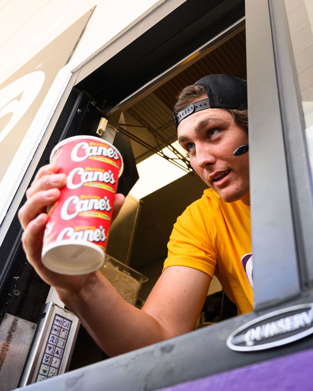 Drive-thru worker in yellow uniform holds stacked Raising Canes cups through service window.