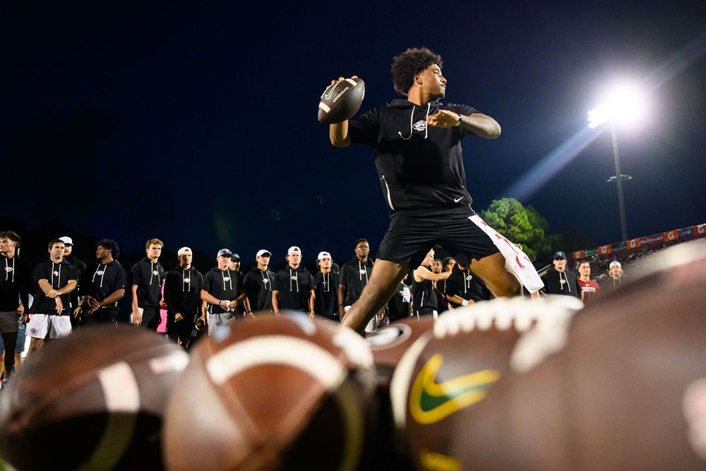 A quarterback practices throwing drills with multiple footballs on the field under stadium lights at night.