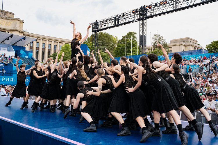 Large group of dancers in black dresses perform synchronized routine on outdoor stage.
