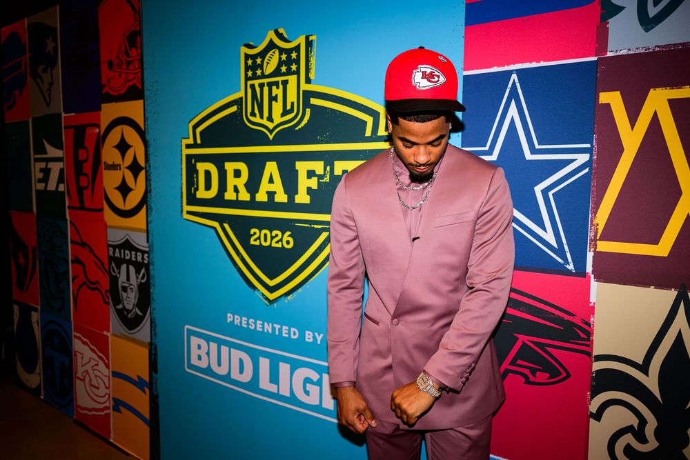 Man in a pink suit and red cap stands before the 2026 NFL Draft backdrop with team logos and a Bud Light sponsor sign.