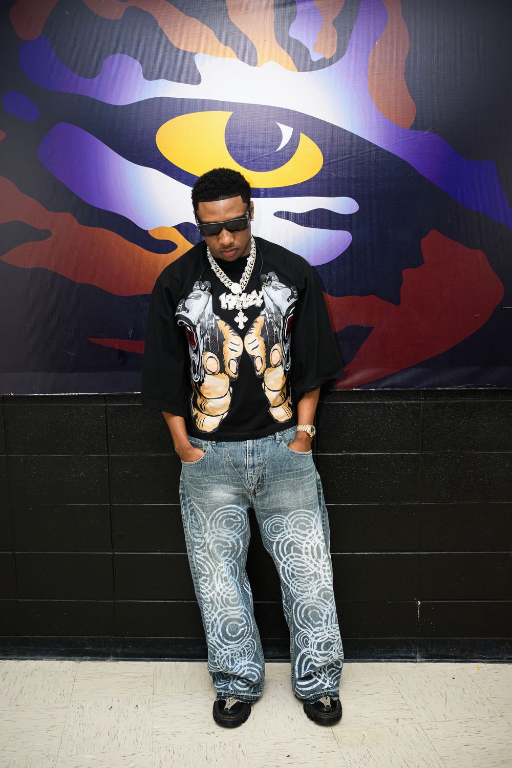 Figure standing against colorful wall mural with large eye design wearing graphic t-shirt and patterned jeans.
