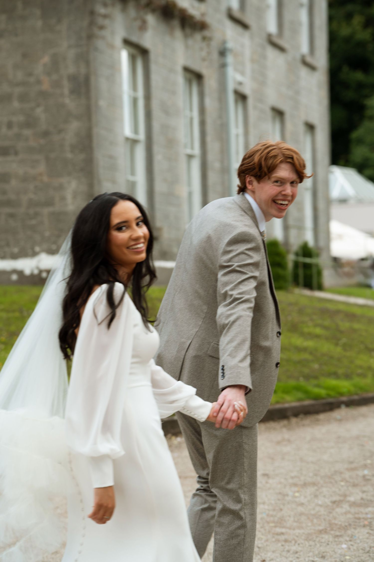 A couple shares playful moments while walking along a stone building pathway on their wedding day.