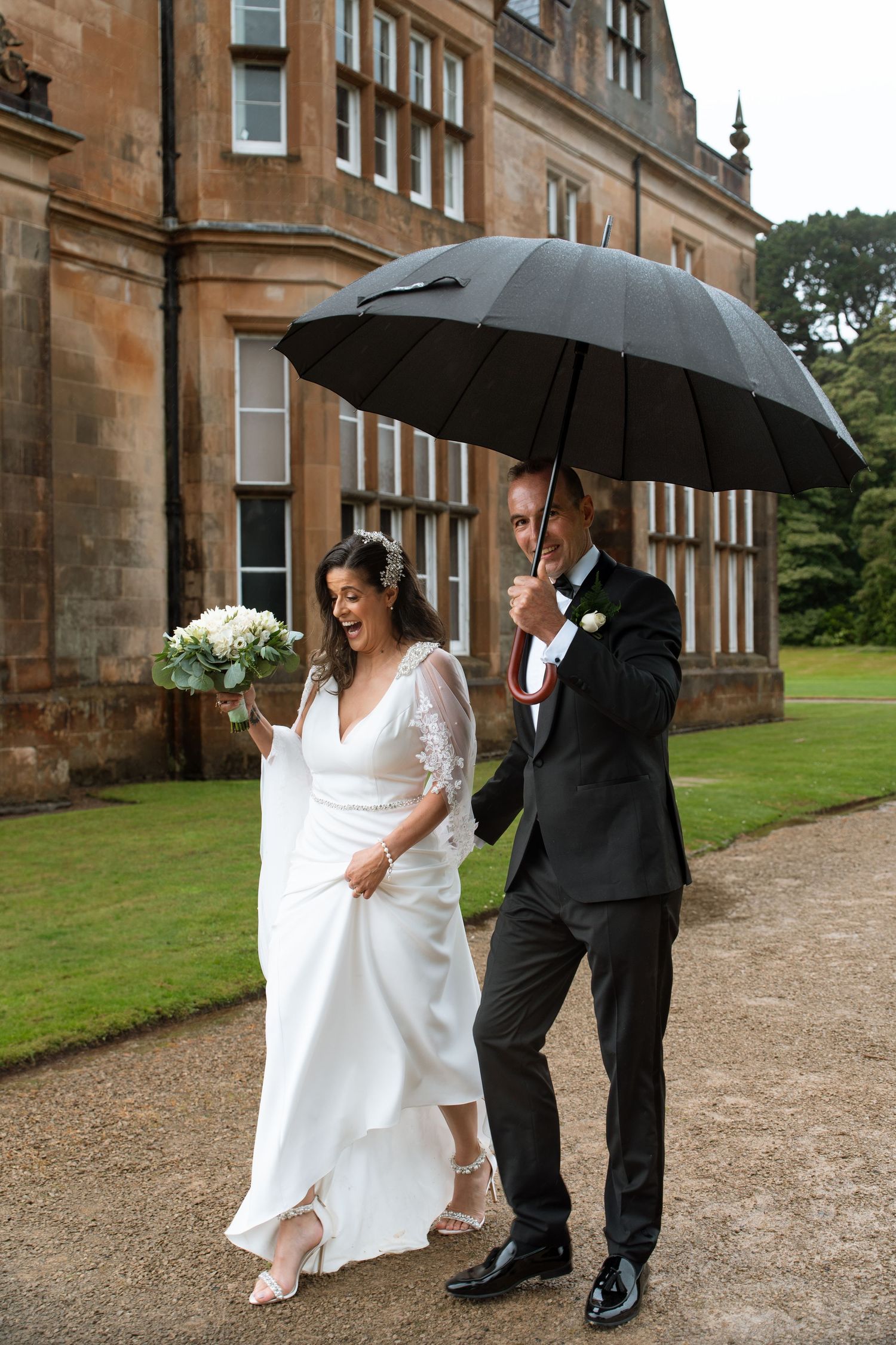 A couple laughs together while sharing an umbrella outside a grand stone building.