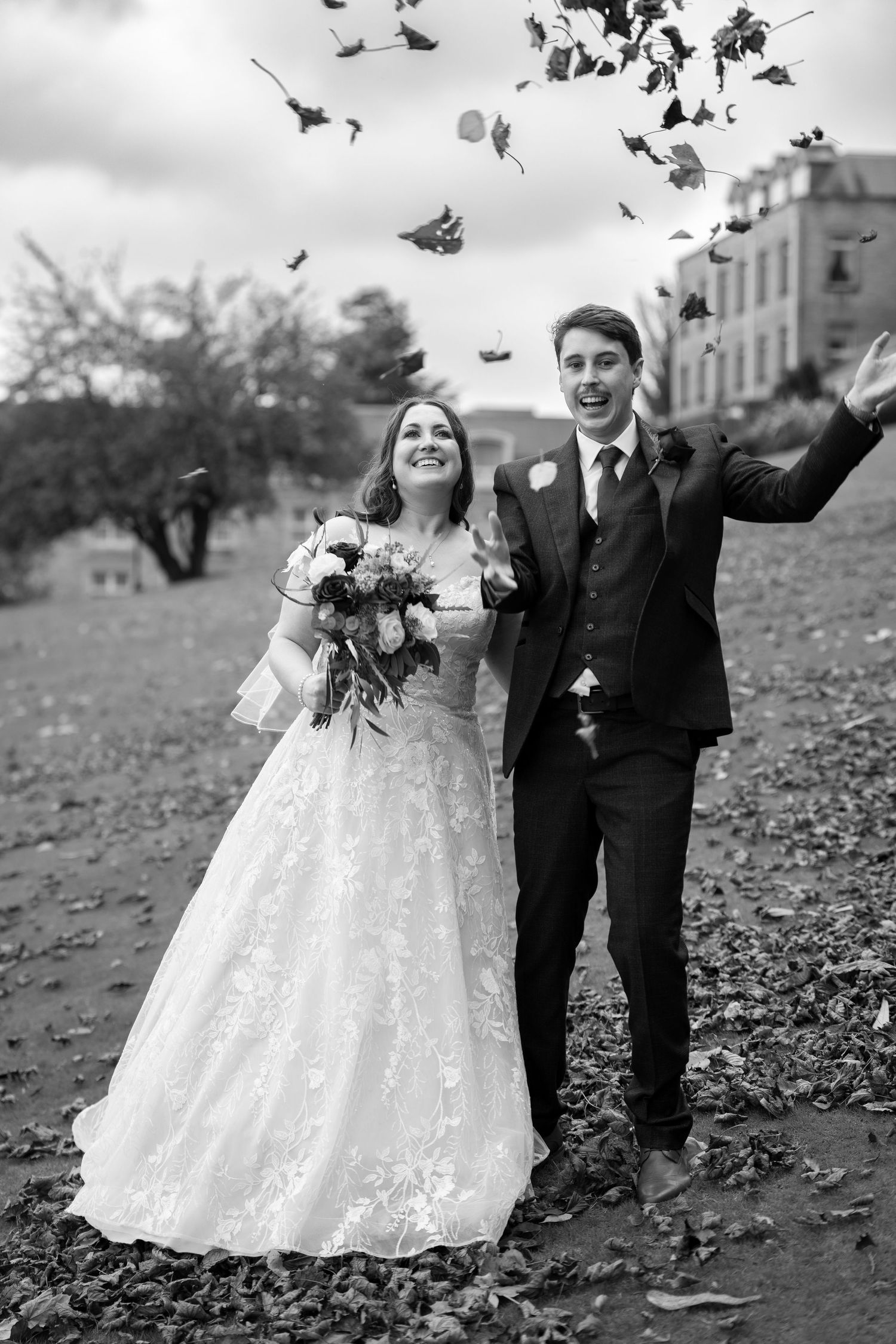 A couple throws autumn leaves in the air during their wedding photoshoot on a cloudy day.
